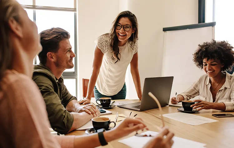 A young woman is standing in front of her computer while collaborating with her colleagues around a work table. 